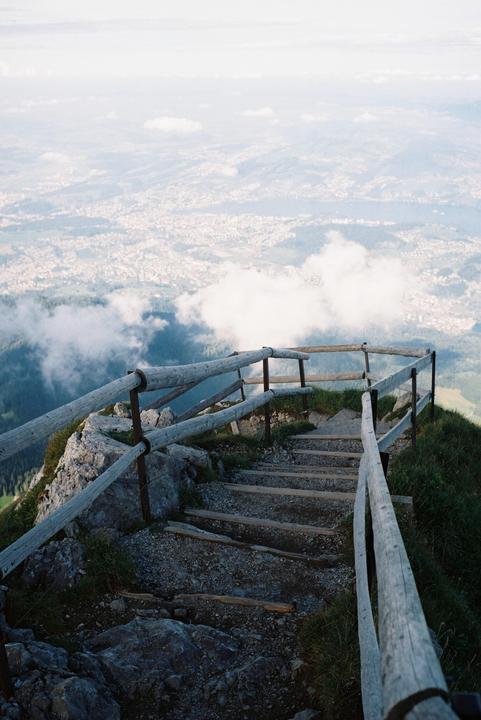 A wooden staircase with a railing descends a rocky mountain path, with a town and clouds visible in the valley below.