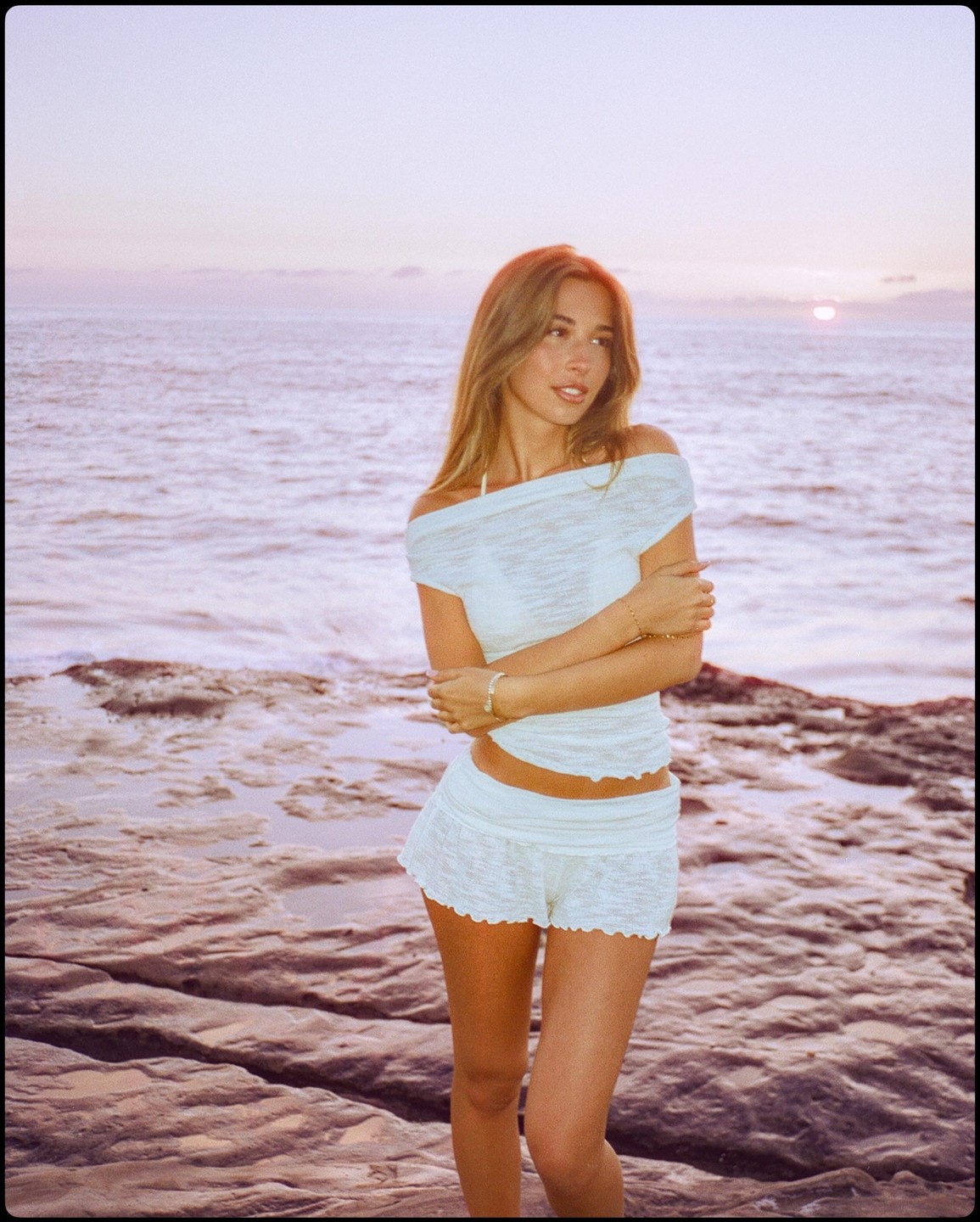 A woman stands on a rocky shore with the ocean and sunset in the background.
