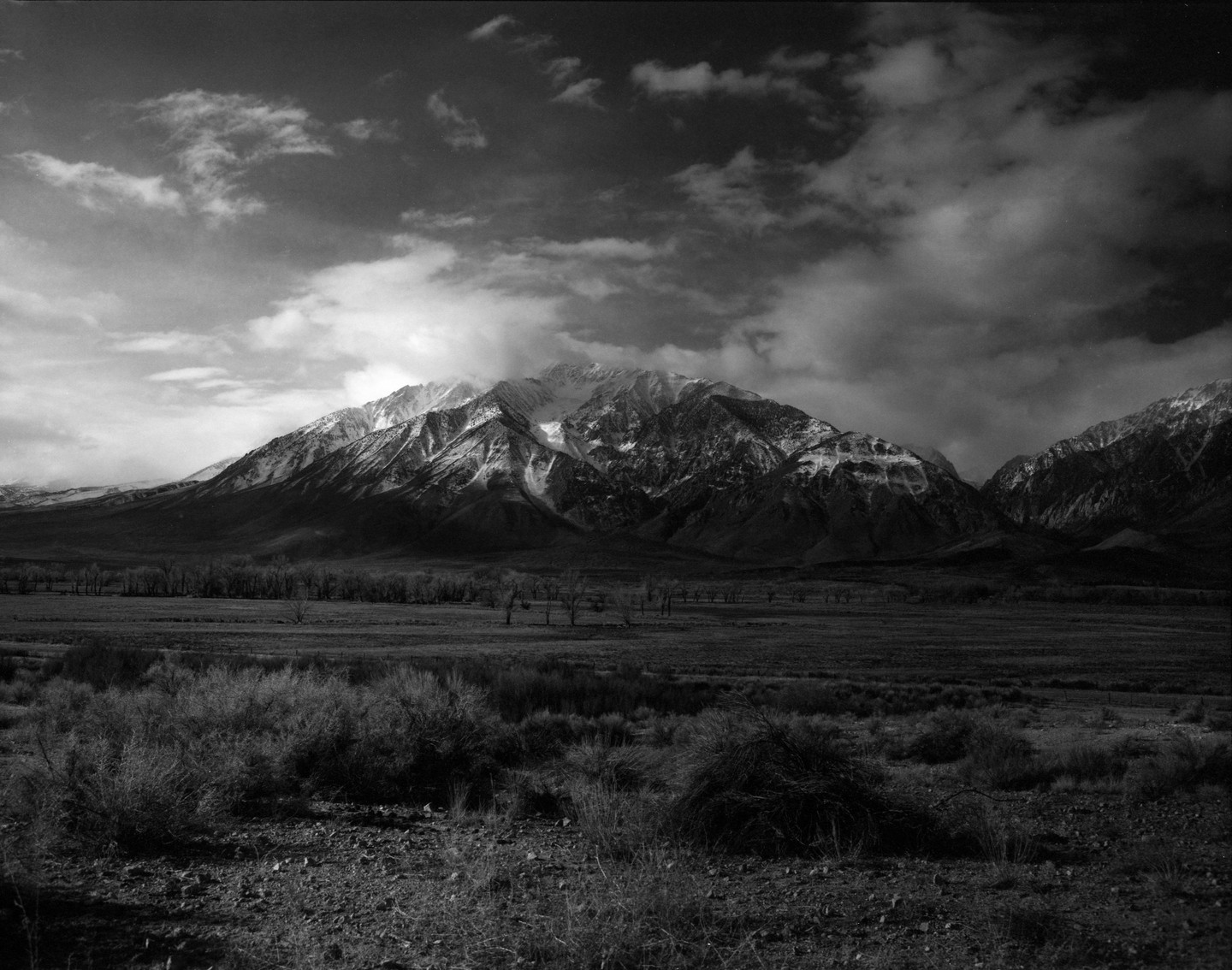 A vast landscape shows a mountain range with snow-capped peaks under a dramatic sky, with a wide-open field in the foreground.