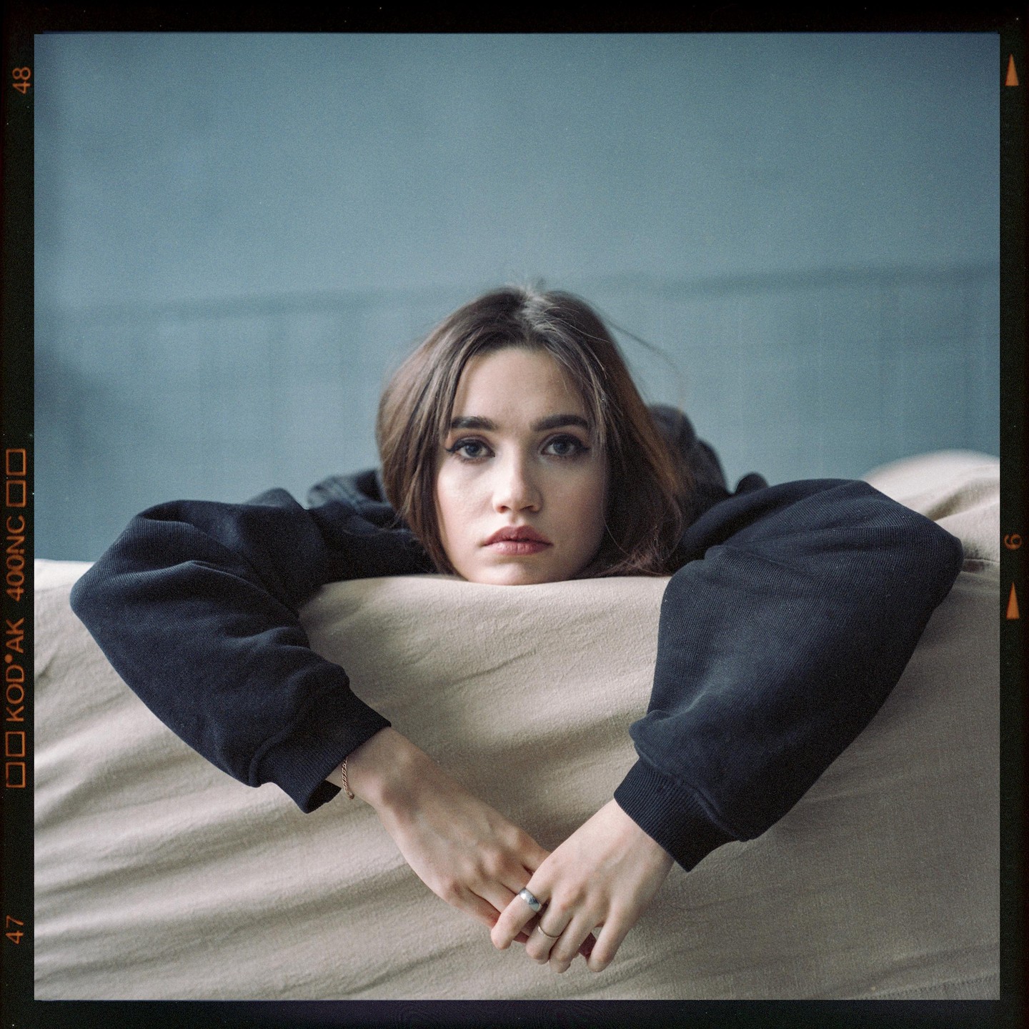 A woman with long hair and a dark sweatshirt rests her head and arms on a light-colored surface.