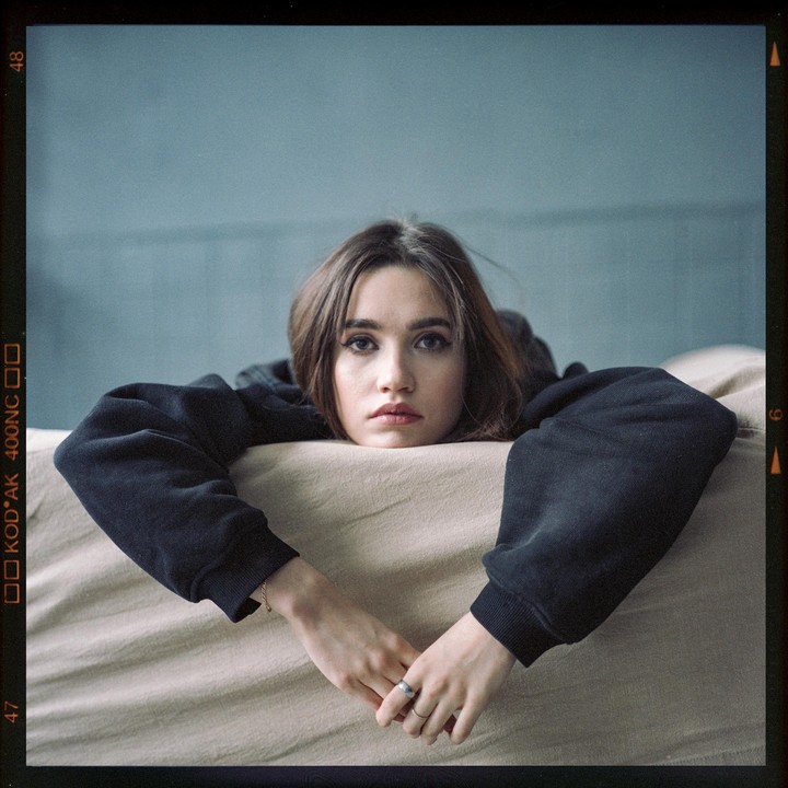 A woman with long hair and a dark sweatshirt rests her head and arms on a light-colored surface.