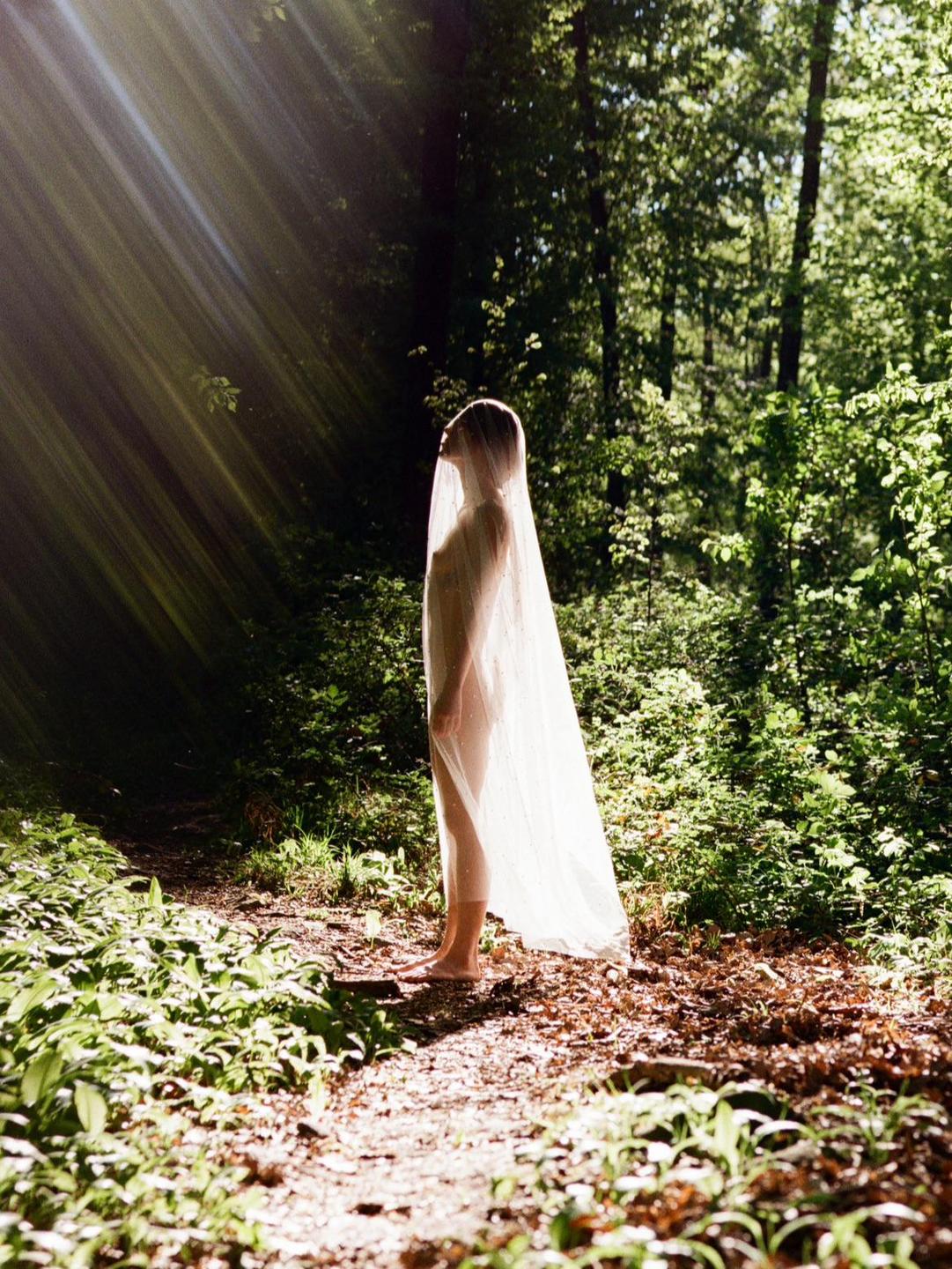 A woman stands barefoot on a forest path, covered by a sheer white veil, with sun rays shining through the trees.