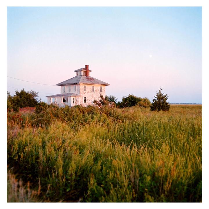 A white house with a red chimney stands in a field of tall grass under a pale sky with a visible moon.