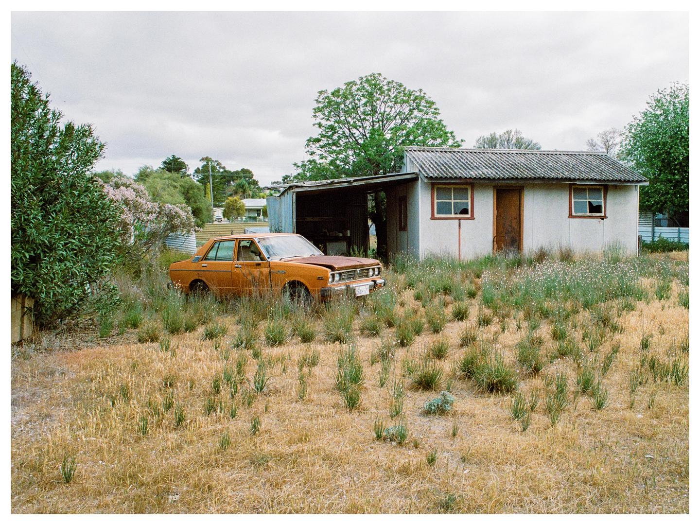An abandoned orange car sits in a grassy field next to a small white building under a cloudy sky.