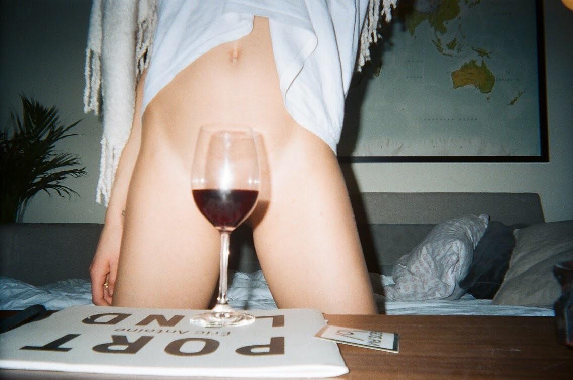 A woman is sitting on a bed with a glass of red wine in front of her, and a world map is on the wall behind her.