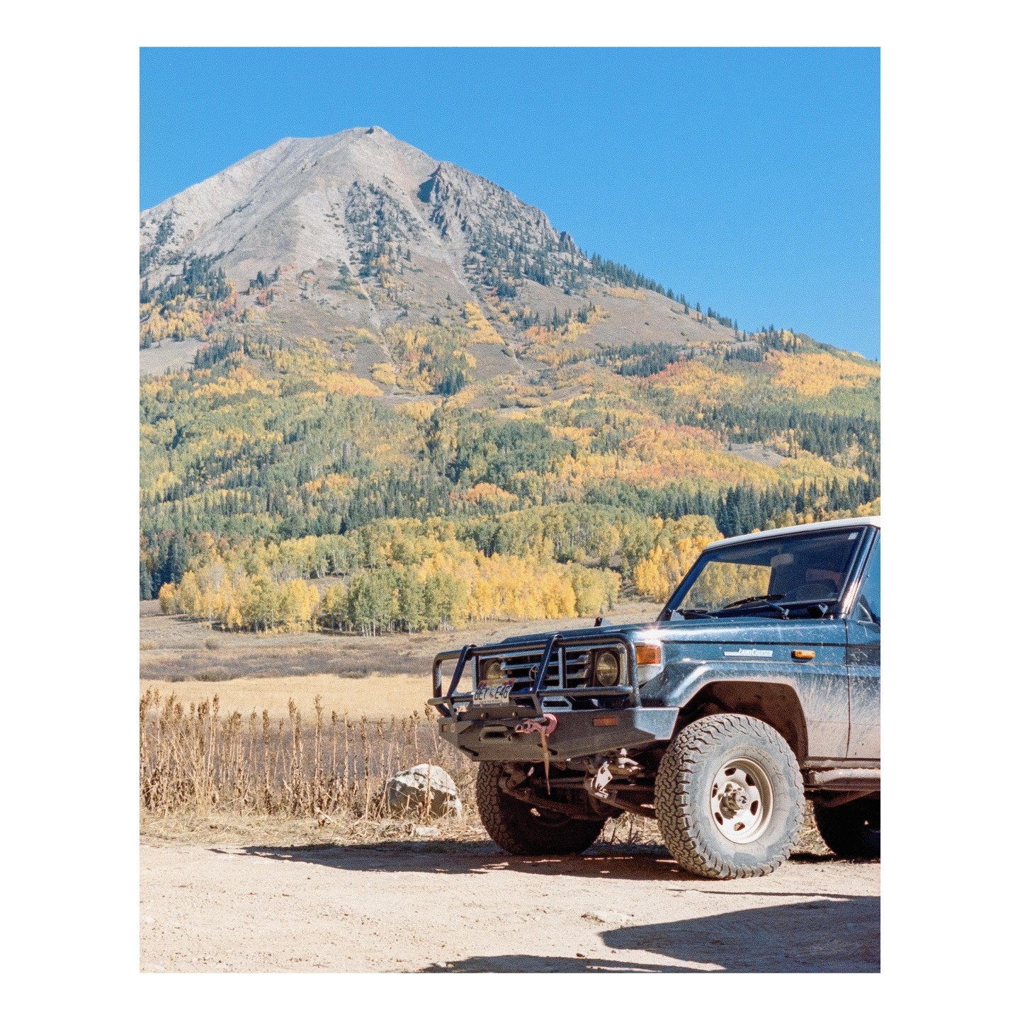 A dark blue off-road vehicle is parked on a dirt road with a mountain and autumn foliage in the background.