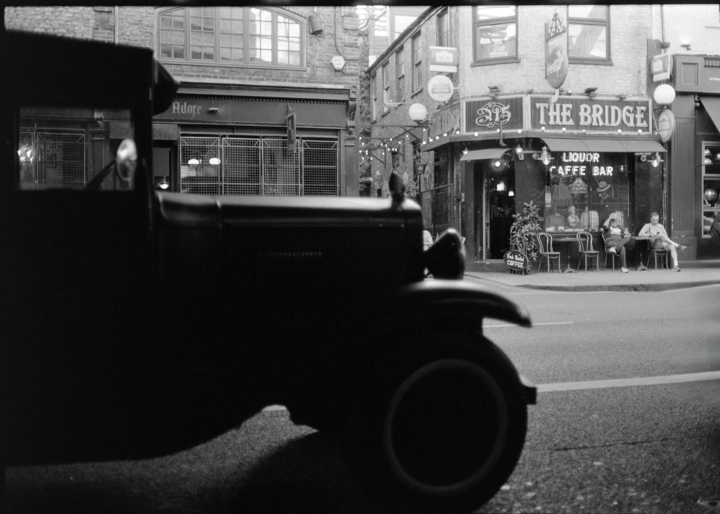A vintage car is parked on a street in front of a bar called The Bridge.