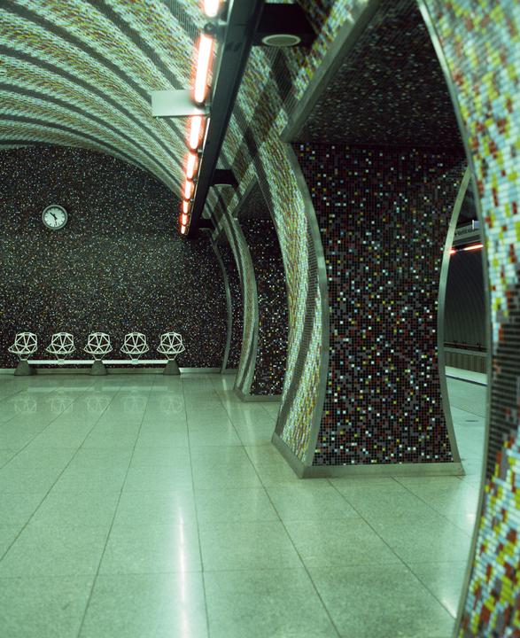 A subway station platform features mosaic-tiled walls and columns, with a row of benches and a clock on the left wall.