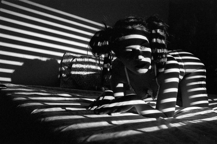A woman with pigtails is on a bed with striped shadows cast across her body and the room.