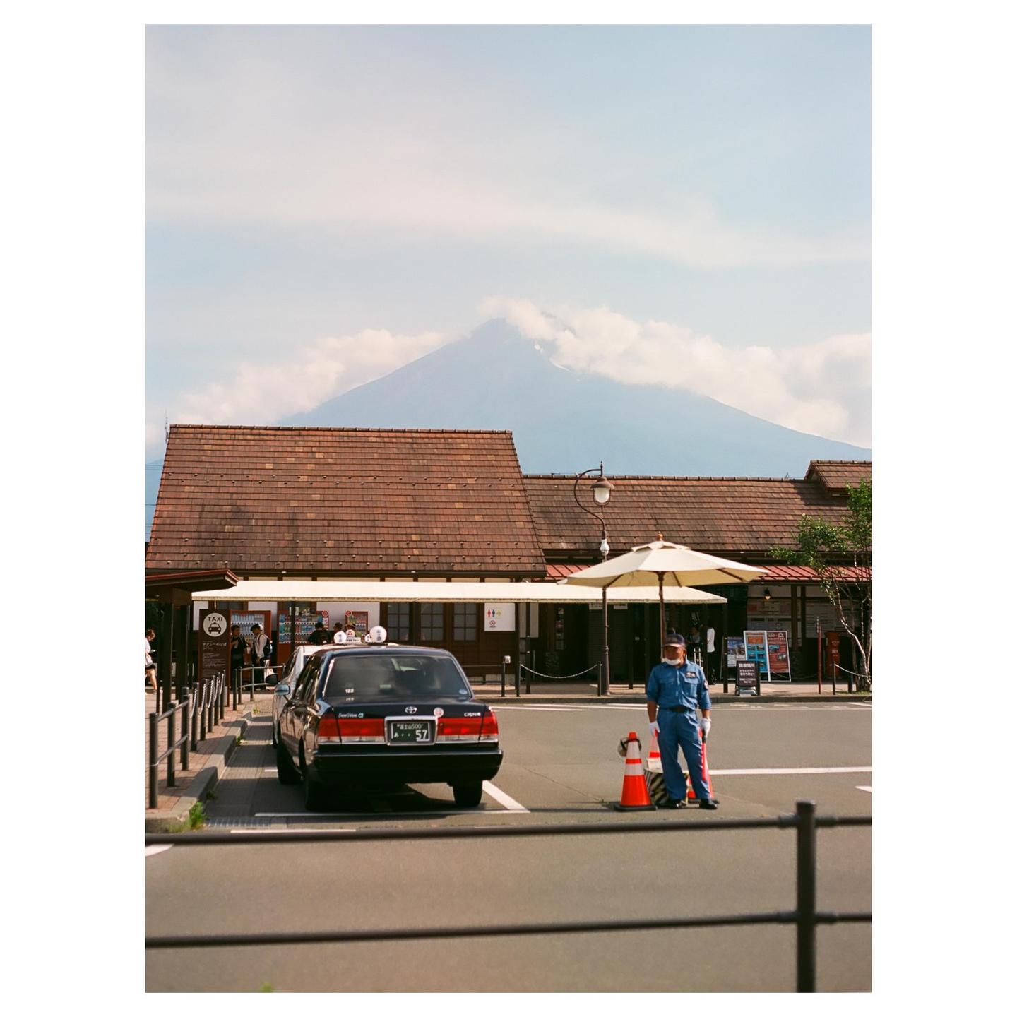 A man in a blue uniform stands next to a taxi in a parking lot with a building and a mountain in the background.