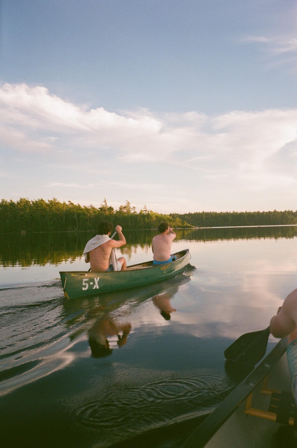 Two men paddle a green canoe across a calm lake surrounded by a forest under a partly cloudy sky.