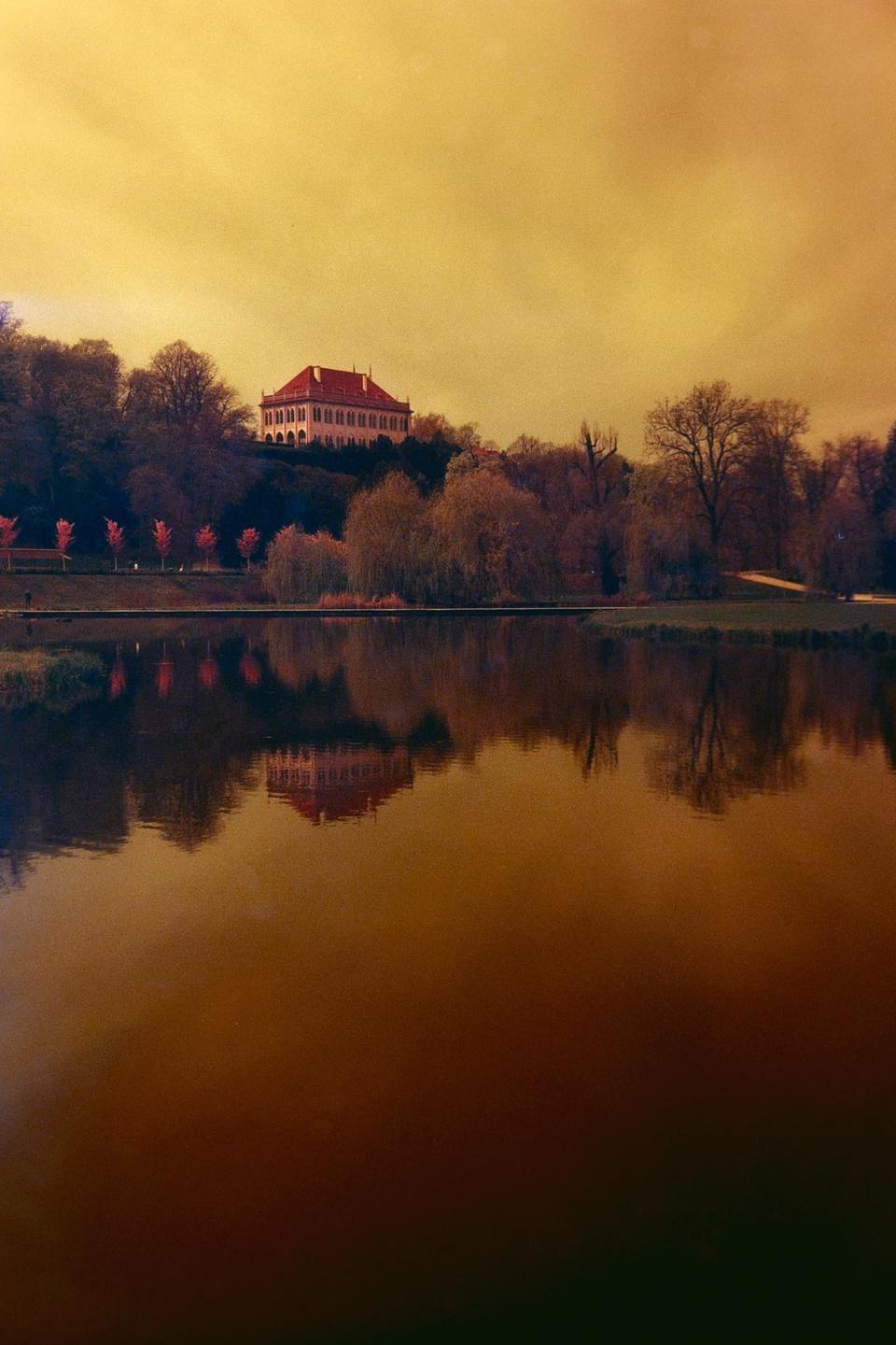 A large building with a red roof sits on a hill overlooking a body of water, reflecting in its surface.