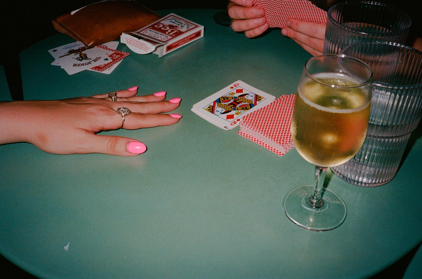 A person's hand with pink nail polish and rings rests on a green table next to playing cards, a glass of champagne, and two empty glasses.