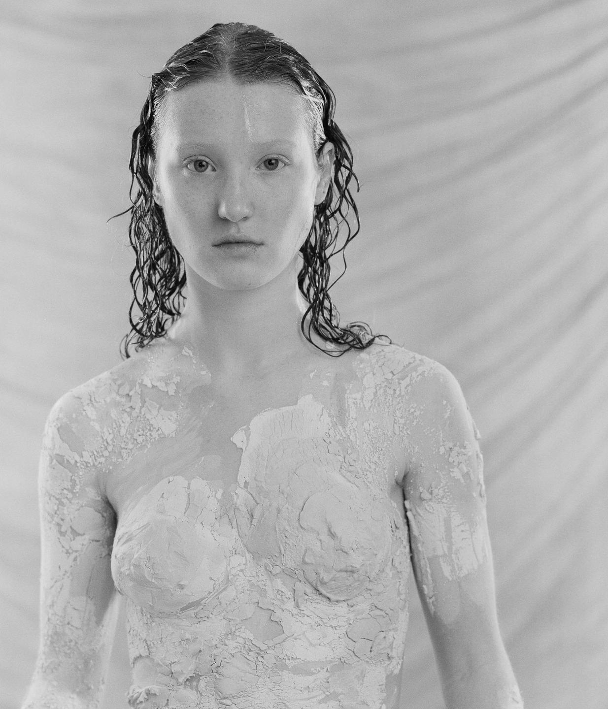 A woman with wet hair and body covered in a crumbling substance looks directly at the viewer.