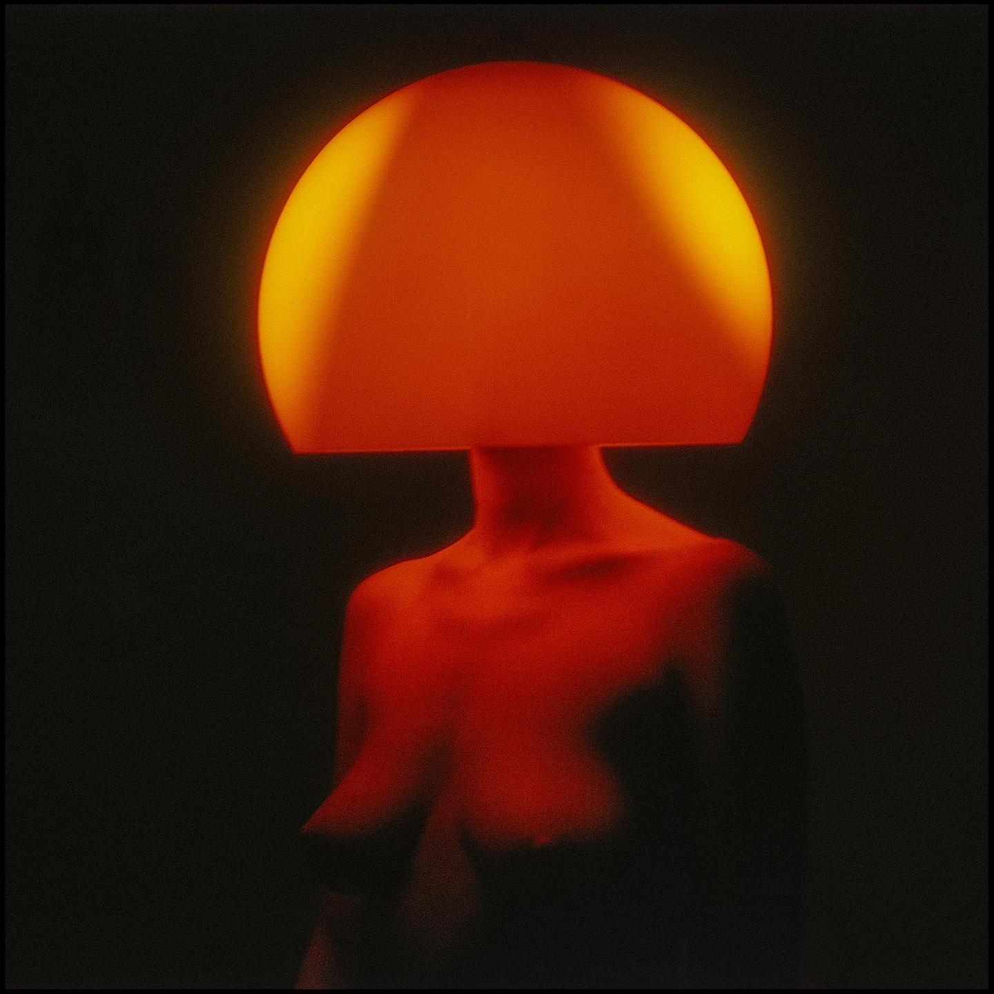 A woman with a glowing orange lampshade for a head stands in the dark.