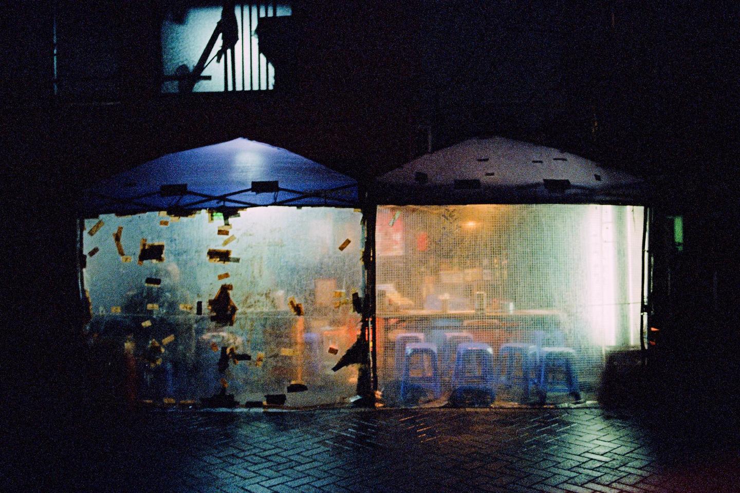 Two makeshift stalls with translucent walls are illuminated from within at night.