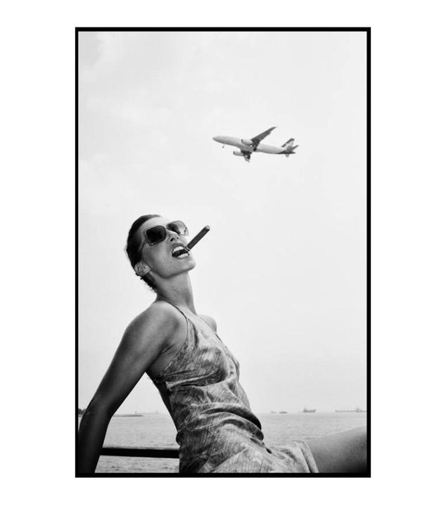 A woman with sunglasses and a cigar in her mouth looks up at an airplane flying overhead.