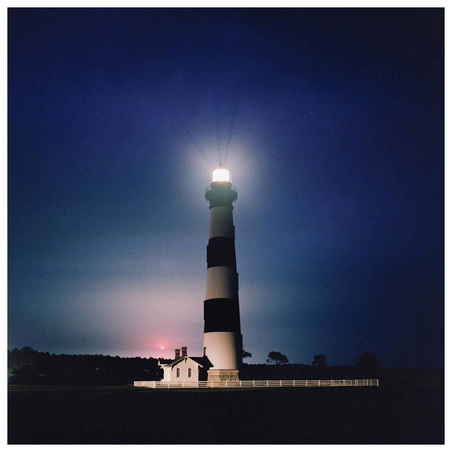 A tall, striped lighthouse shines brightly against a dark blue night sky, with a small white building and fence at its base.