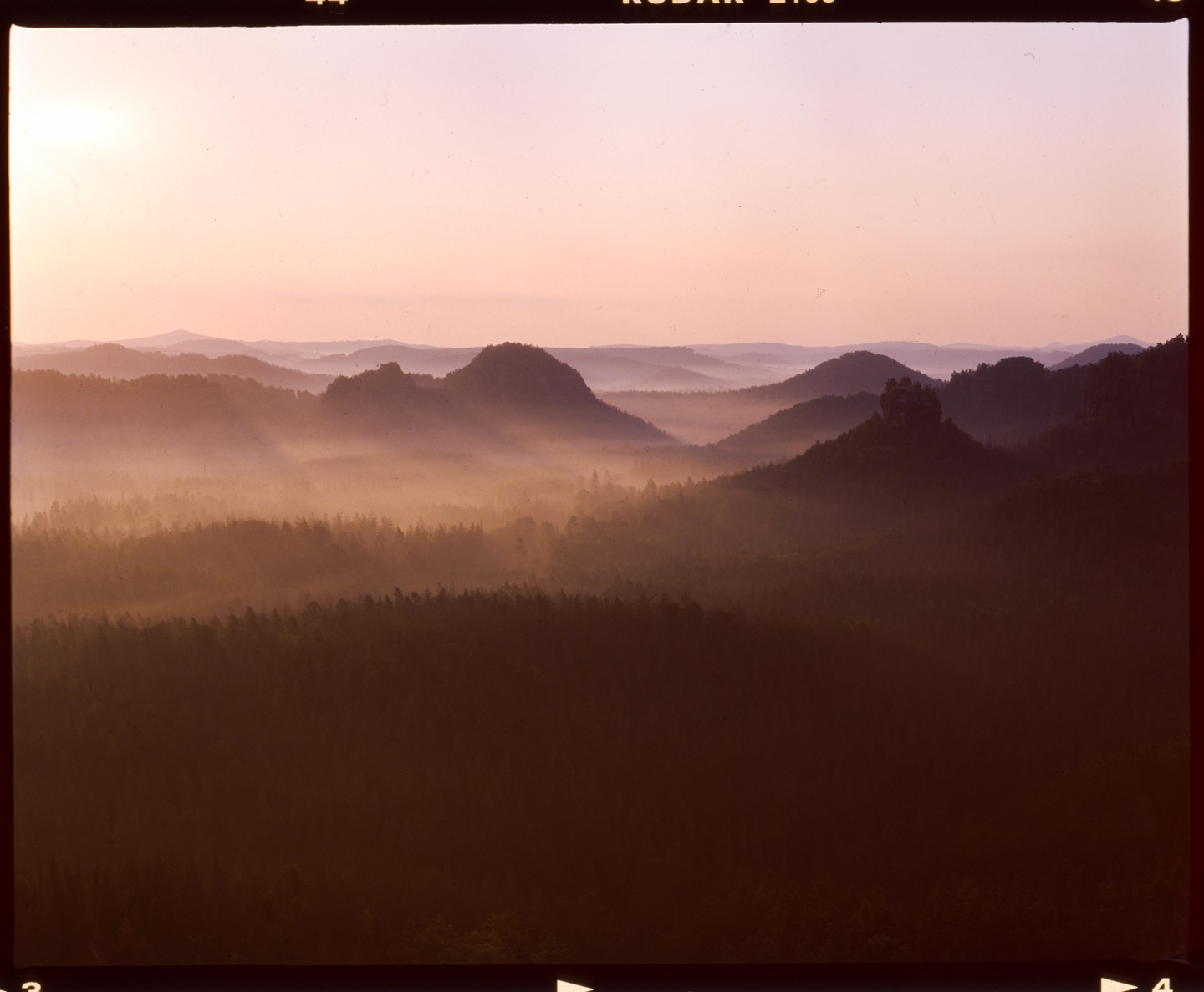 A hazy landscape shows layers of mountains and forests under a pale sky.
