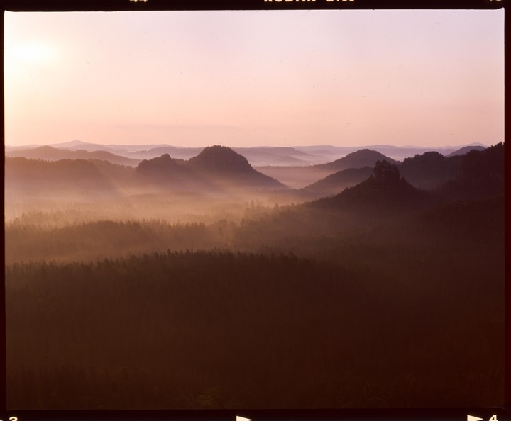 A hazy landscape shows layers of mountains and forests under a pale sky.