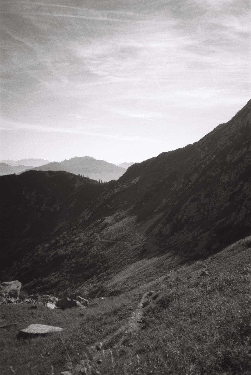 A winding path leads up a grassy mountain slope with distant peaks under a cloudy sky.