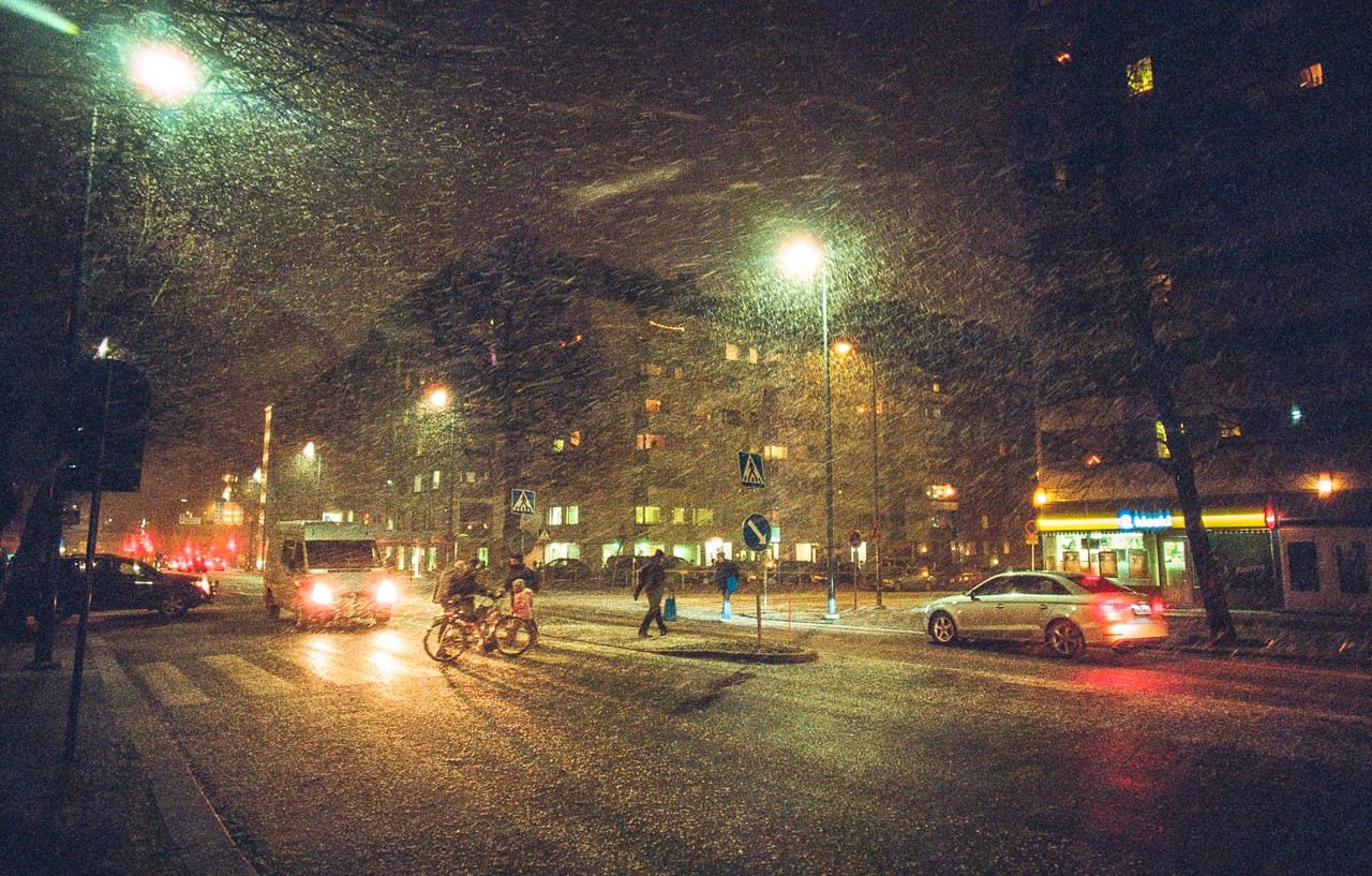 A snowy street at night shows cars, people, and a cyclist passing by buildings with illuminated windows.
