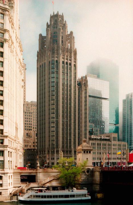 A tall, ornate building with a flag on top stands prominently among other city buildings, with a boat visible on the water below.