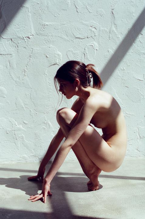 A woman crouches on a floor next to a textured wall with shadows.