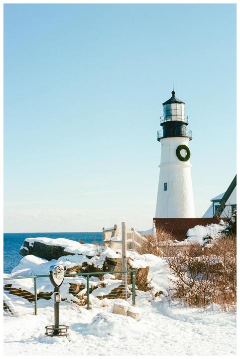 A white lighthouse with a green wreath on its side stands on a snowy cliff overlooking the ocean under a clear blue sky.