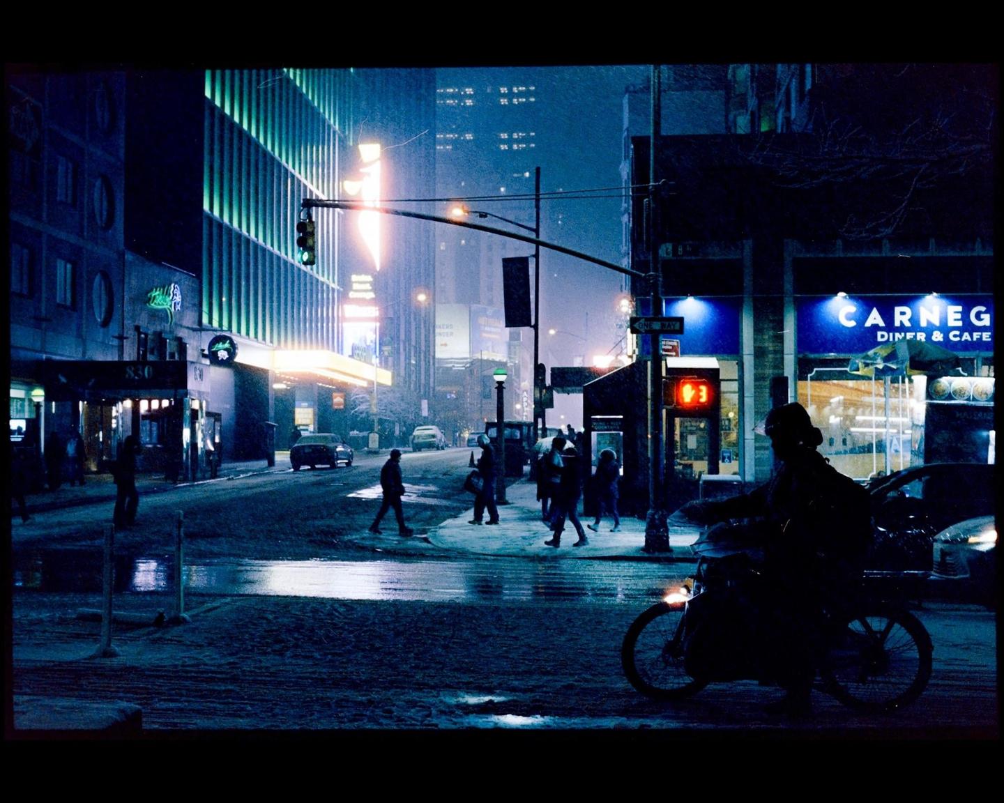 A street scene at night shows people walking and a person on a bicycle, with illuminated buildings and falling snow.