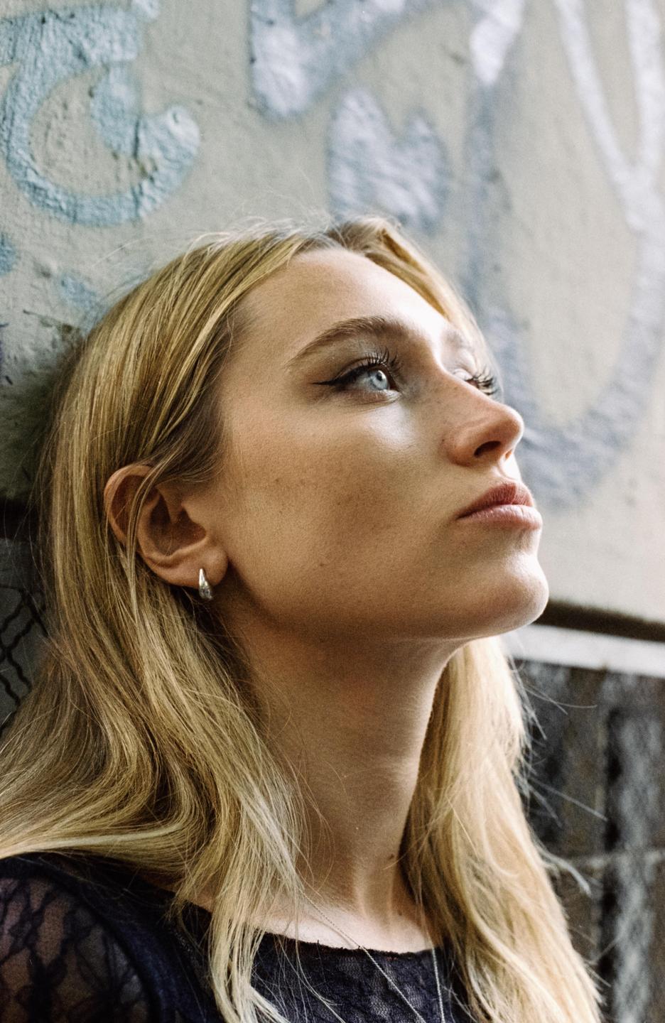 A woman with long blonde hair and blue eyes looks up while leaning against a graffiti-covered wall.