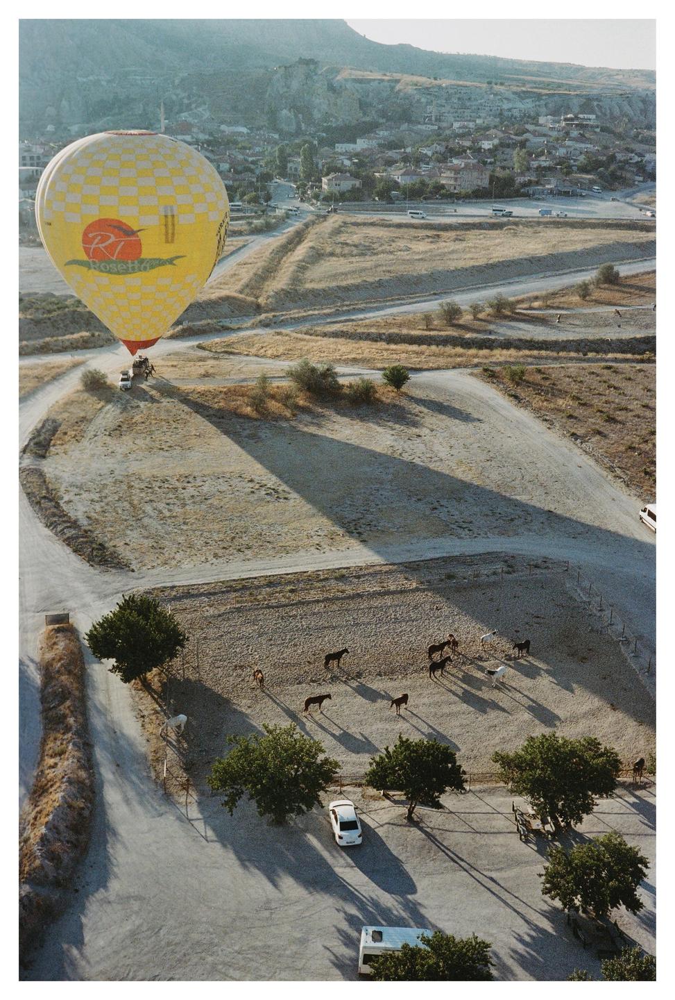 A yellow hot air balloon casts a long shadow over a dry landscape with horses and a distant town.