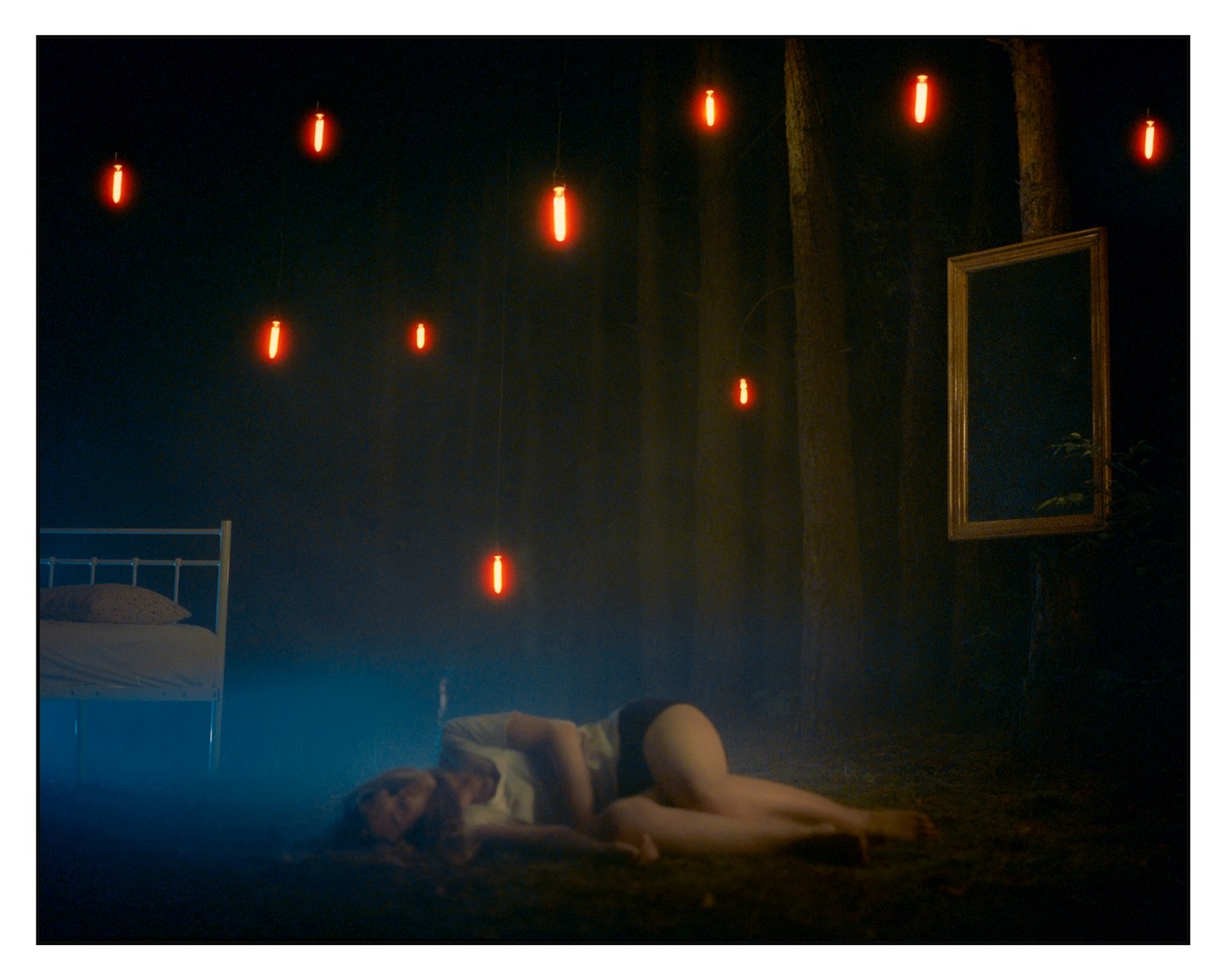A woman lies on the ground in a dark forest with a bed, a mirror, and glowing red lights hanging from trees.