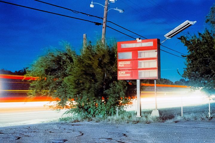 A red gas price sign stands beside a road with light trails from passing vehicles at night.
