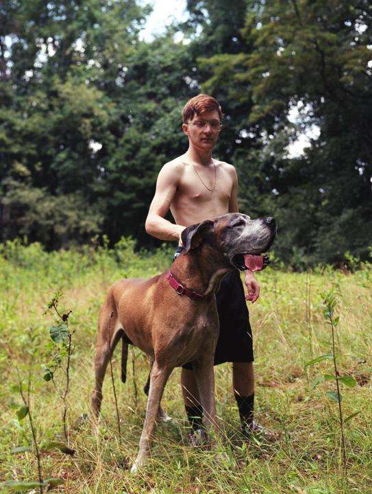 A man stands shirtless in a field with a large dog looking up at him.