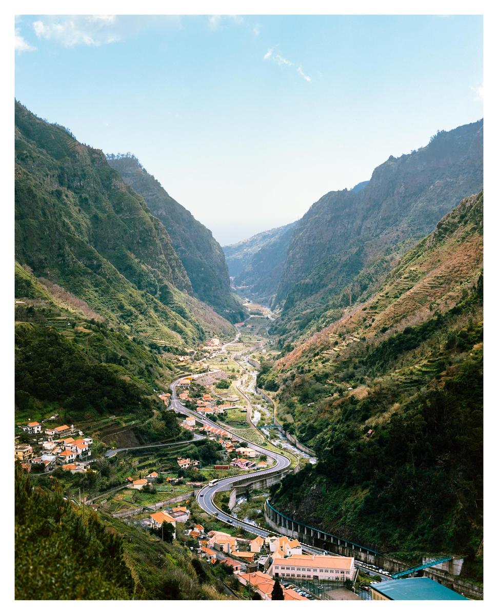 A winding road and a small village are nestled in a deep valley surrounded by lush green mountains under a clear sky.