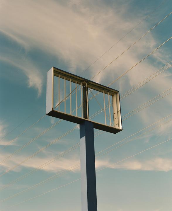 An empty billboard stands against a blue sky with wispy clouds and power lines.