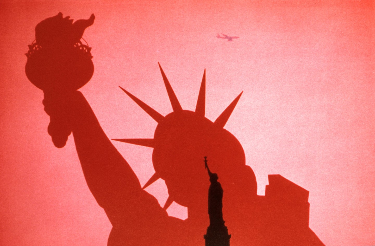 A double exposure shows a silhouette of the Statue of Liberty in front of a red sky with an airplane and a smaller Statue of Liberty.