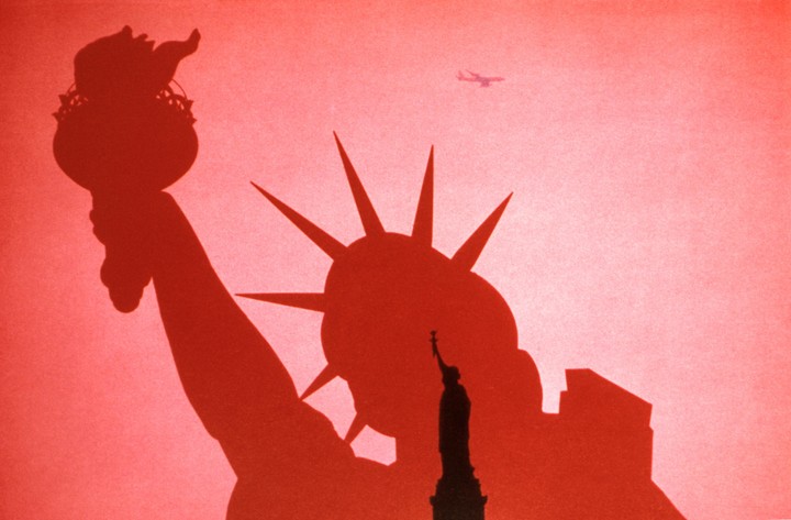 A double exposure shows a silhouette of the Statue of Liberty in front of a red sky with an airplane and a smaller Statue of Liberty.