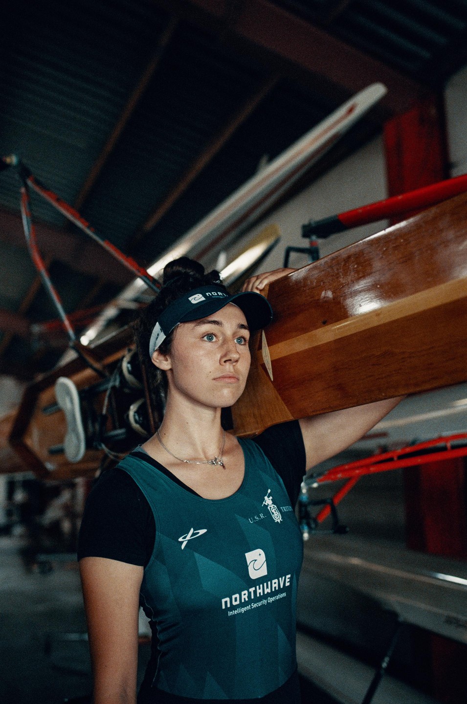 A woman in athletic wear carries a wooden rowing shell on her shoulder inside a boathouse.
