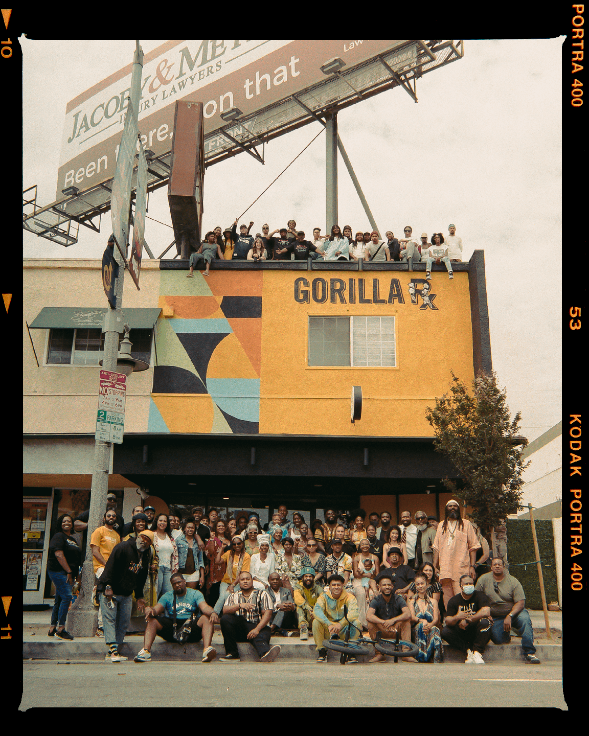 A large group of people are gathered in front of a yellow building with a billboard above it.