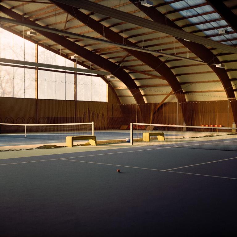 A tennis ball rests on a blue indoor tennis court with two nets and benches under a high arched ceiling.