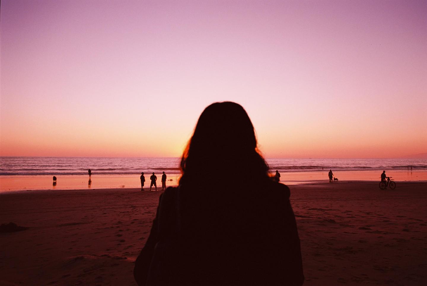 A person with long hair stands on a beach looking out at the ocean and sunset with other people in the distance.