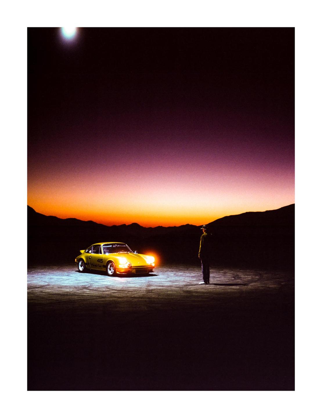 A man stands next to a gold car with its headlights on, against a sunset sky and mountains.
