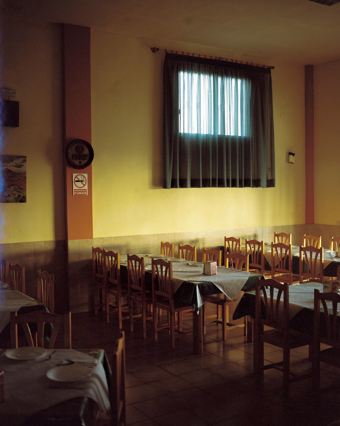 A restaurant dining room with many tables and chairs is illuminated by light from a window.