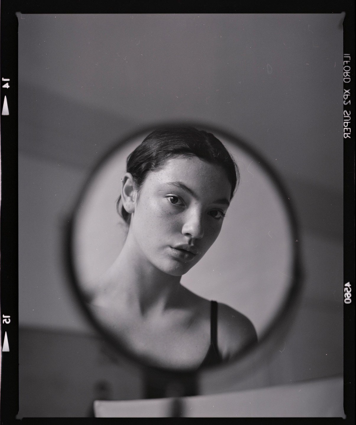 A woman's face is reflected in a round mirror, with her shoulders and chest also visible.