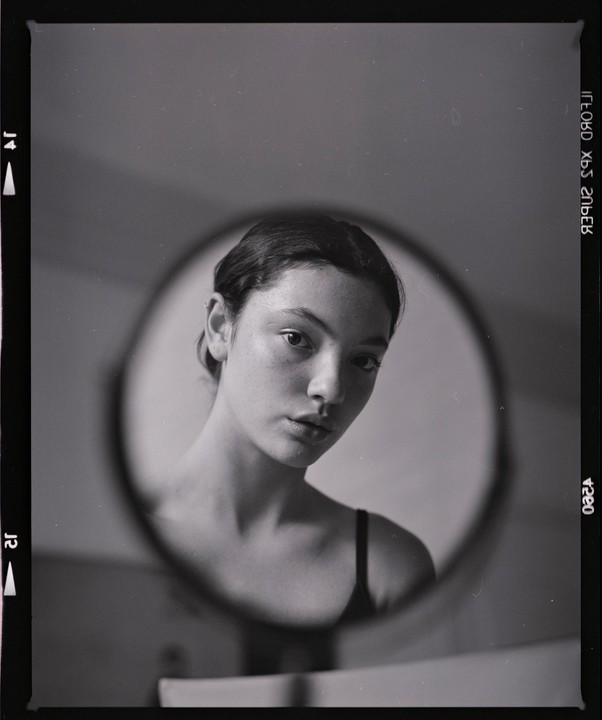 A woman's face is reflected in a round mirror, with her shoulders and chest also visible.