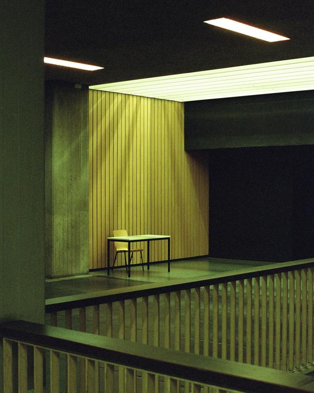 A table and two chairs sit in a room with wood paneling and concrete walls, viewed from above a railing.