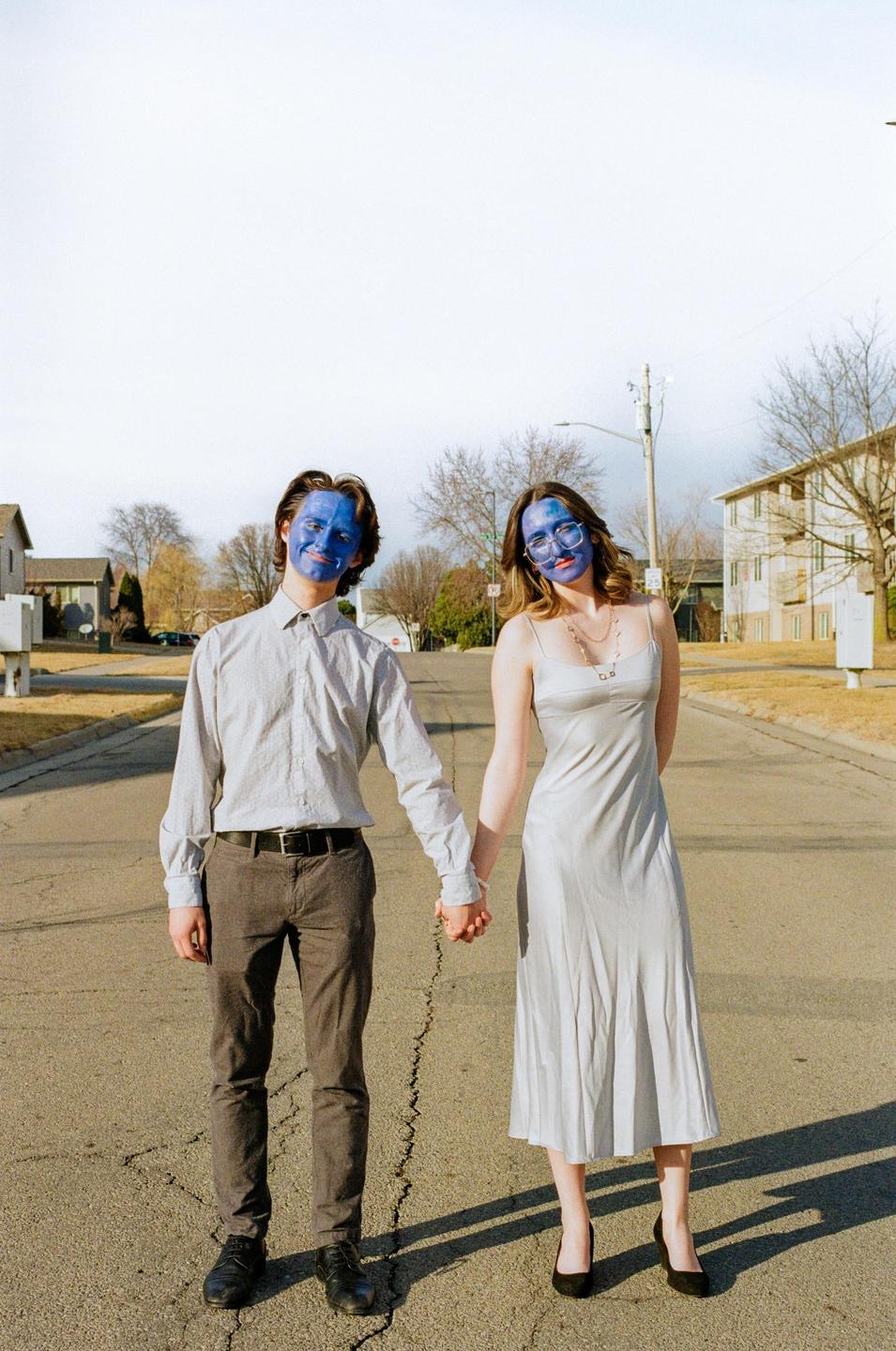 A couple with blue painted faces stands holding hands in the middle of a cracked street.