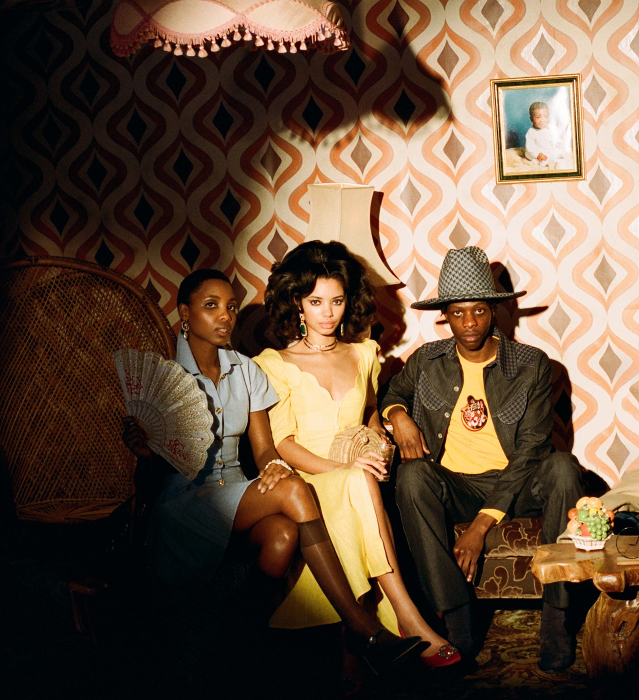 Three people sit on a sofa in a room with patterned wallpaper and a framed picture on the wall.